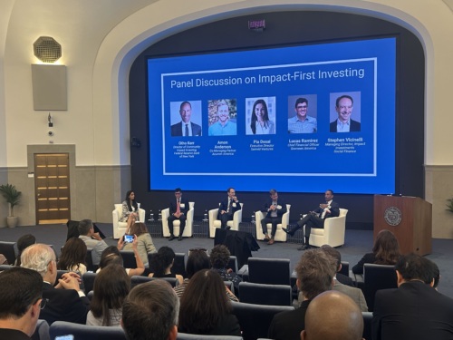 Five panelists sit onstage in front of a large screen titled “Panel Discussion on Impact-First Investing” as an audience observes and takes photos in a modern auditorium.