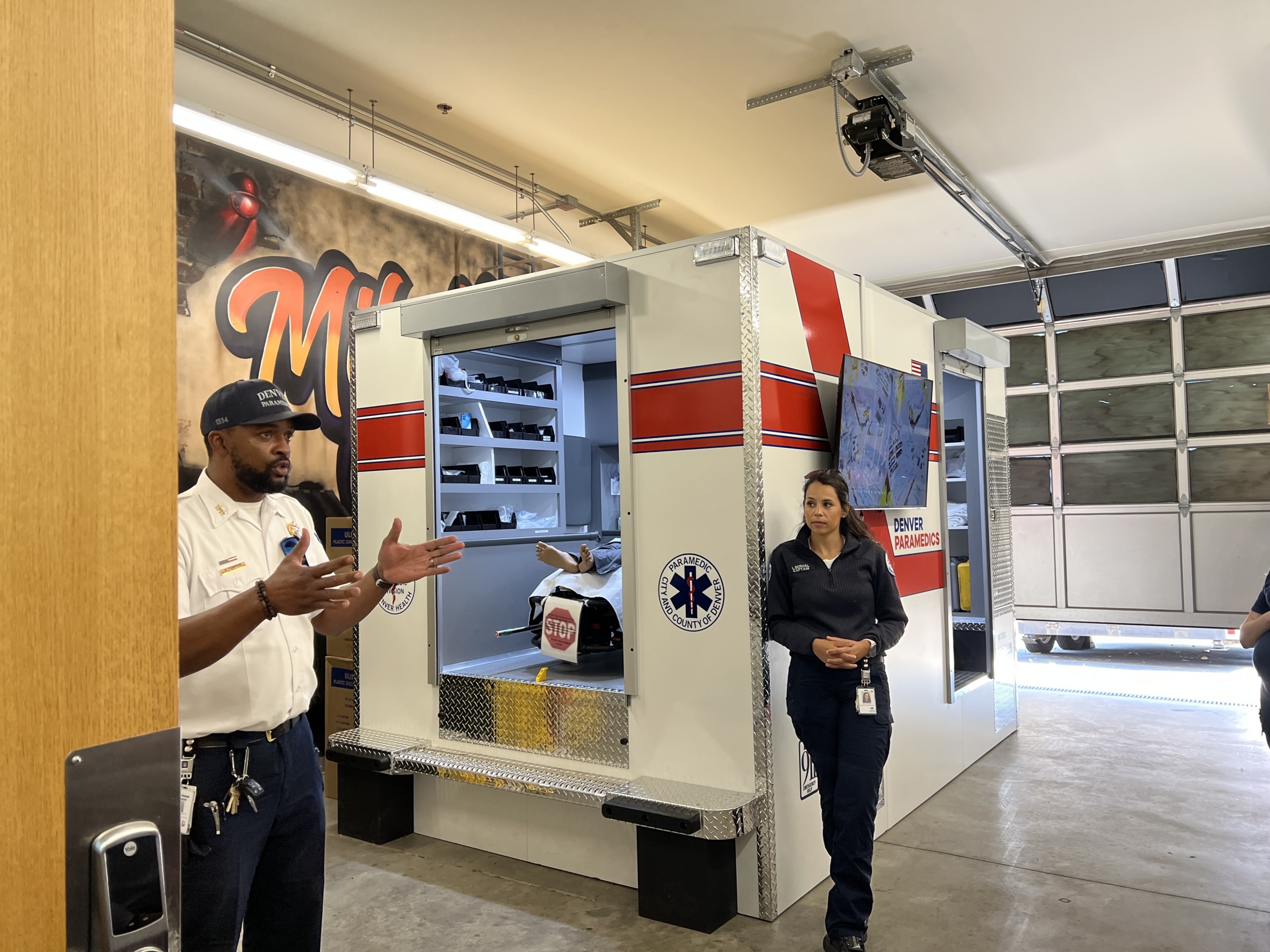 Two people stand next to a medical supply unit inside a garage. The man gestures as he speaks, while the woman listens. Shelves and a monitor displaying “DRONE HANGAR” are visible.