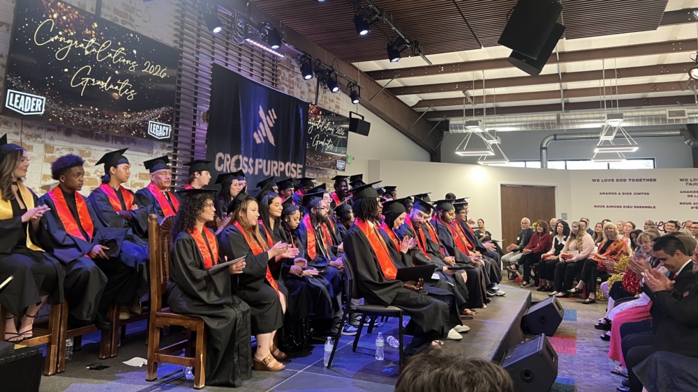 A group of graduates in caps and gowns sit on stage facing an audience in a modern indoor venue during a graduation ceremony.