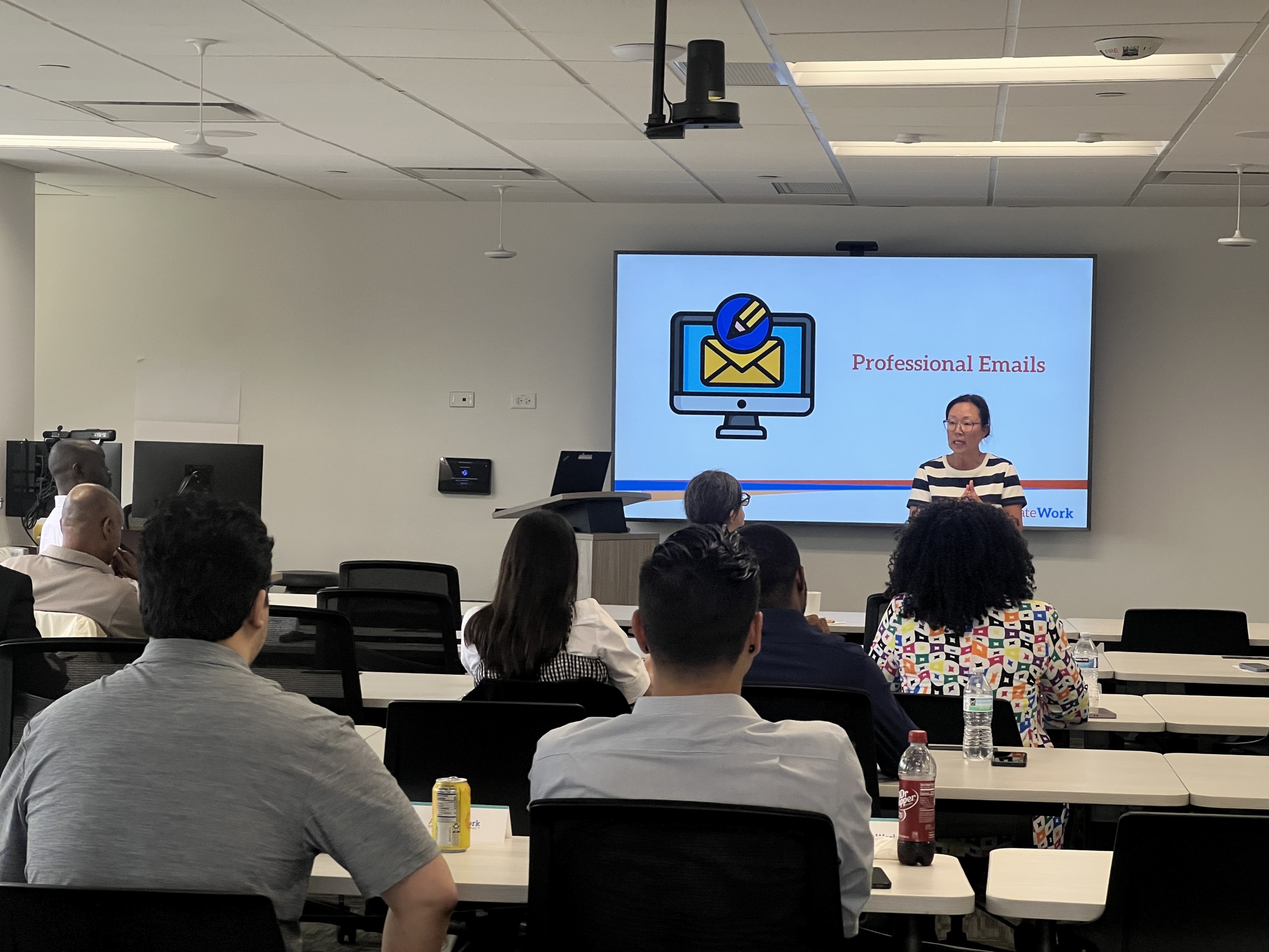 A person stands at the front of a classroom giving a presentation titled “Professional Emails” to seated attendees.