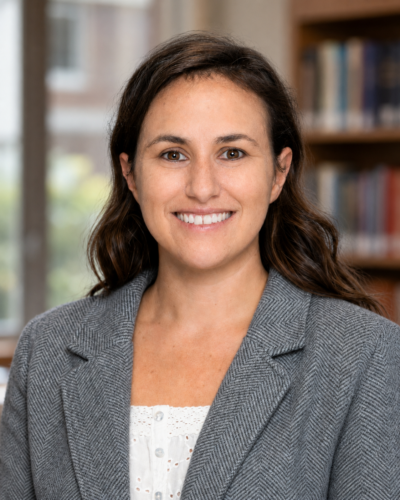 Woman with long brown hair wearing a gray blazer and white top, smiling, standing in a library or office setting.