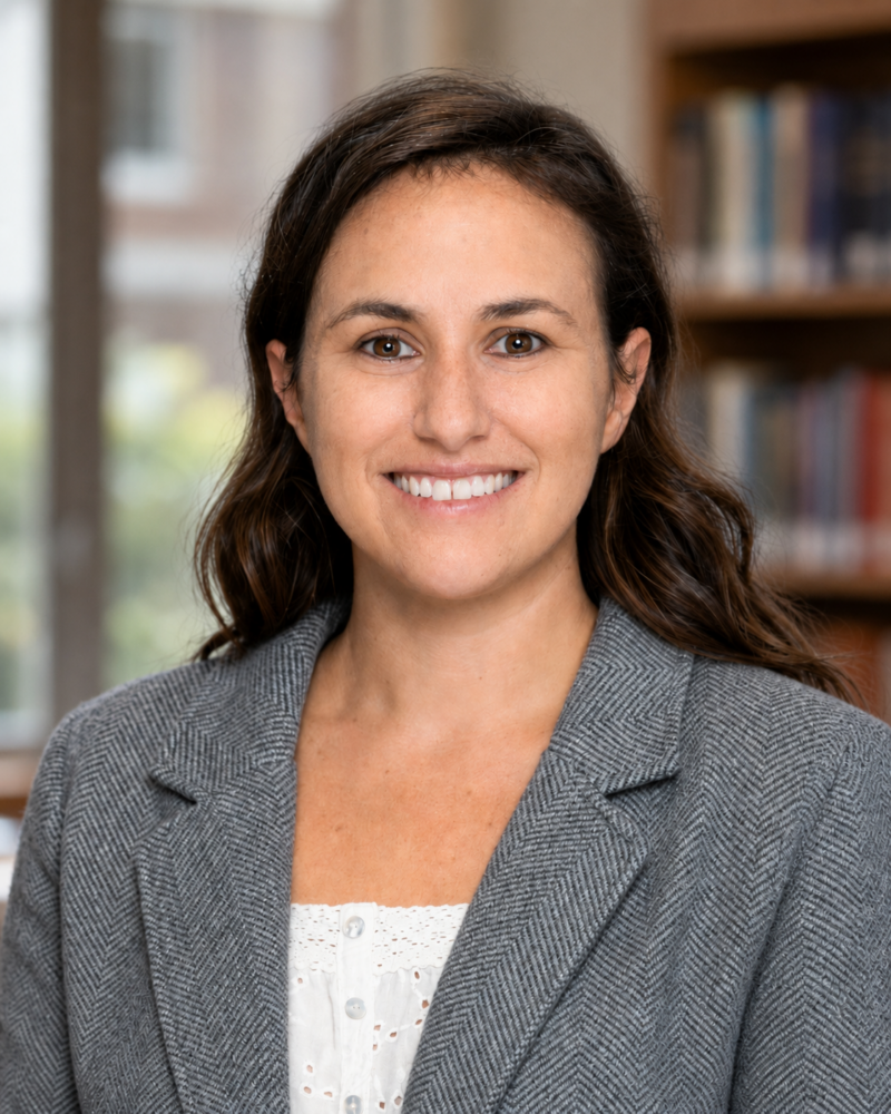 Woman with long brown hair wearing a gray blazer and white top, smiling, standing in a library or office setting.
