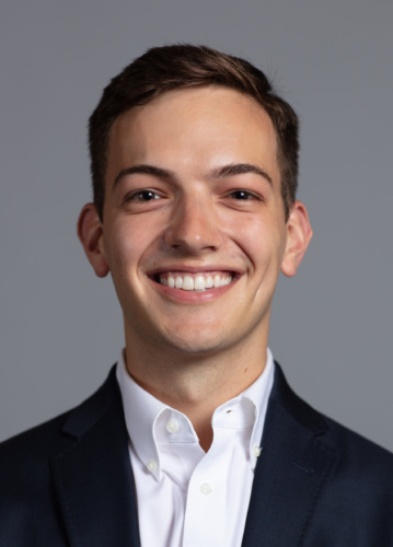 A young man with short brown hair, wearing a dark suit jacket and white button-down shirt, smiling at the camera against a plain gray background.