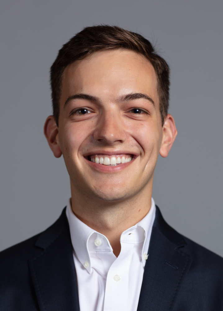 A young man with short brown hair, wearing a dark suit jacket and white button-down shirt, smiling at the camera against a plain gray background.