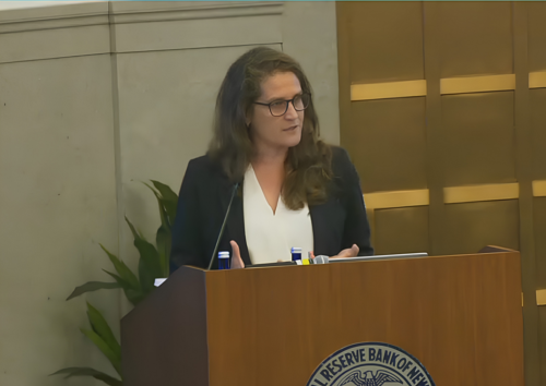 A woman with long hair and glasses speaks at a podium with the Reserve Bank of New Zealand emblem, standing indoors near a plant and a wall with wooden panels.