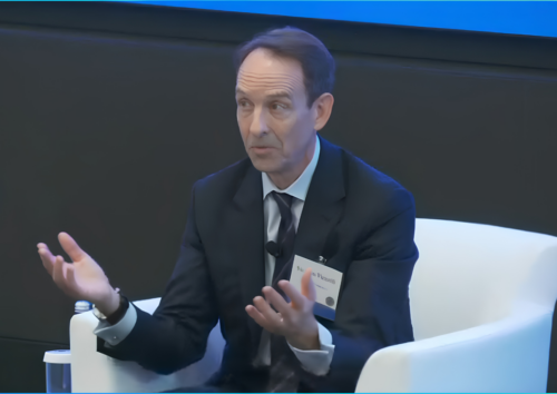 A man in a suit sits in a white chair, speaking with hands raised. He has a name tag, and a glass of water is on a table beside him. A blue screen is in the background.