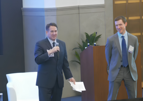 Two men in suits stand at the front of a room; one speaks into a microphone while holding papers, the other stands near a podium and smiles.