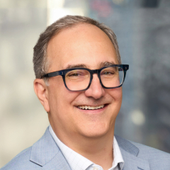 Smiling middle-aged man wearing glasses and a light blue blazer, standing in front of a blurred background.