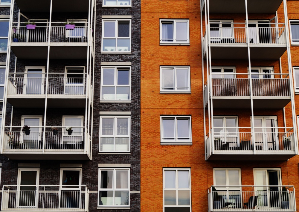 Exterior view of a modern apartment building with orange and dark brick walls, balconies with railings, and several visible potted plants.