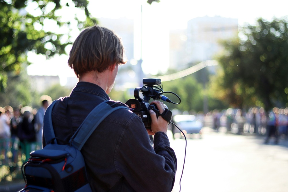 A person with a backpack films a scene outdoors using a professional video camera.