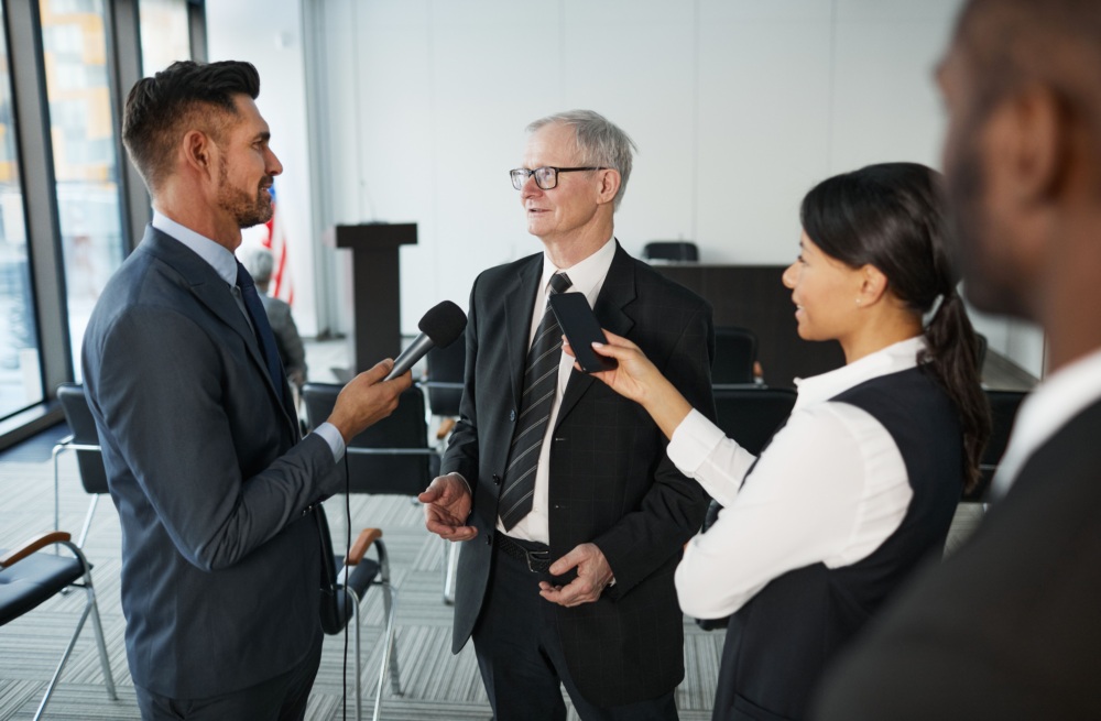 A businessman in a suit speaking into a microphone held by a reporter, with another person recording him with a smartphone.